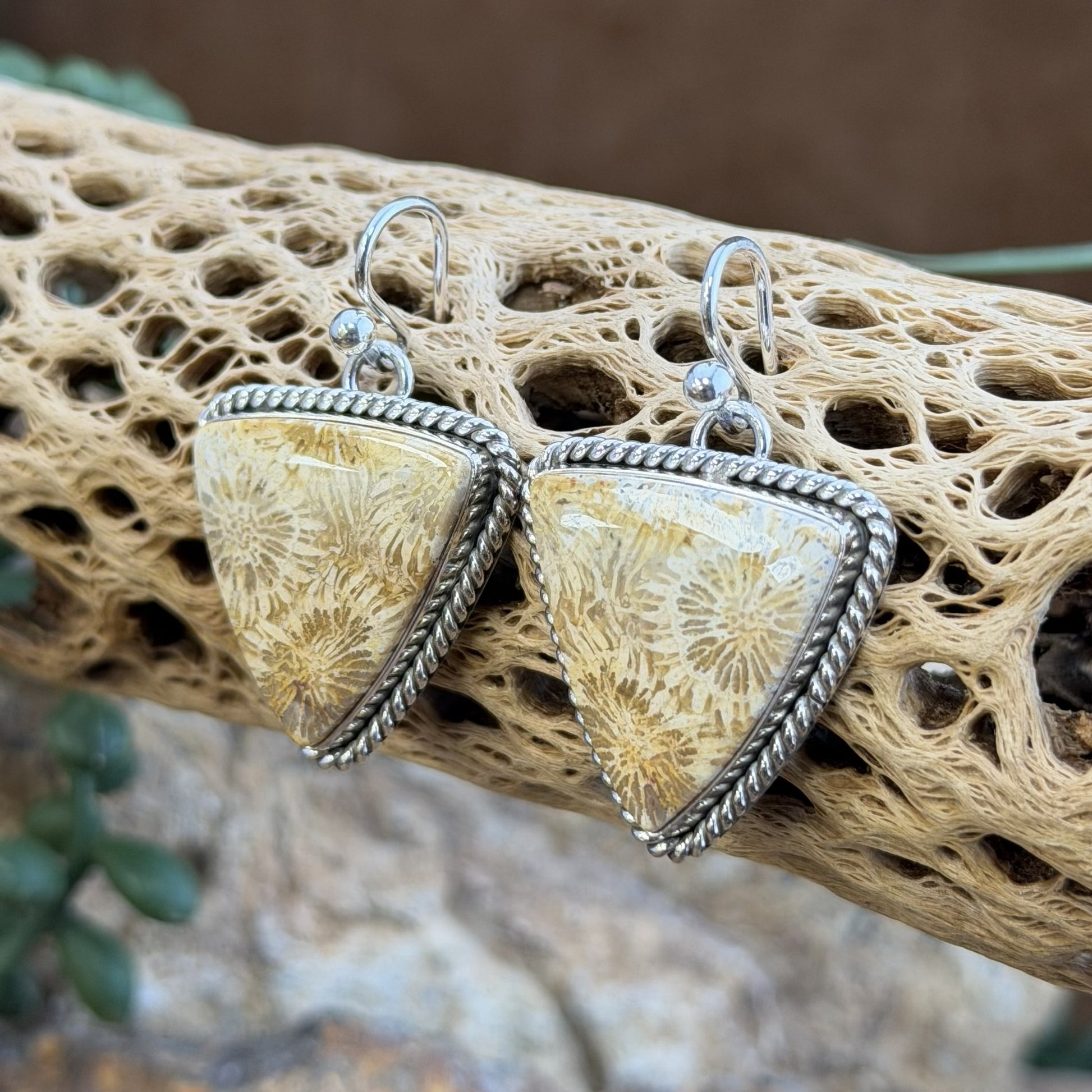 A pair of triangular dangle earrings with textured beige and cream flower like patterns set in silver-toned twisted rope borders and fishhook wires, displayed on a natural driftwood branch.