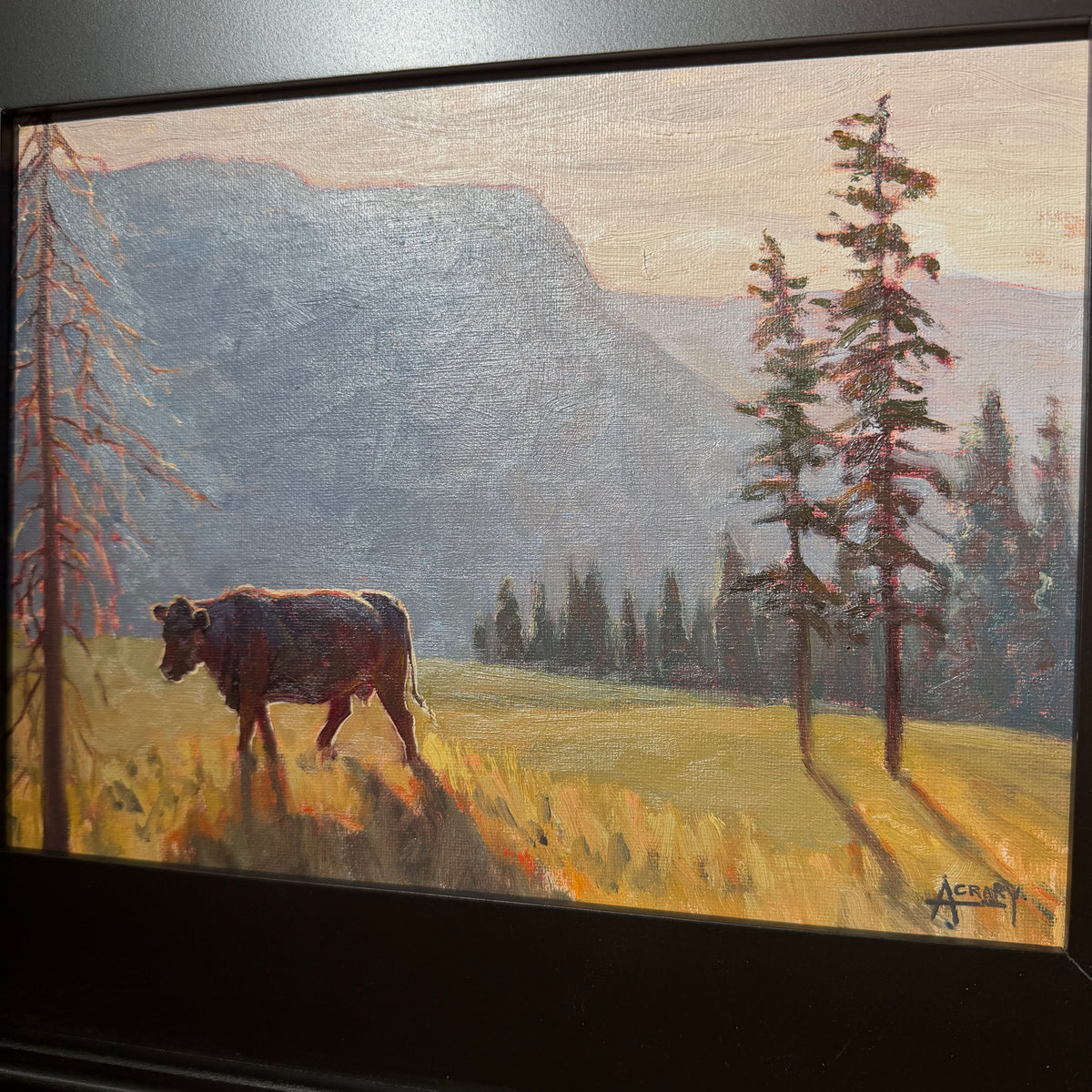 A framed landscape painting depicting a cow standing in a sunlit grassy field with tall trees and blue-gray mountains in the background.