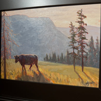 A framed landscape painting depicting a cow standing in a sunlit grassy field with tall trees and blue-gray mountains in the background.