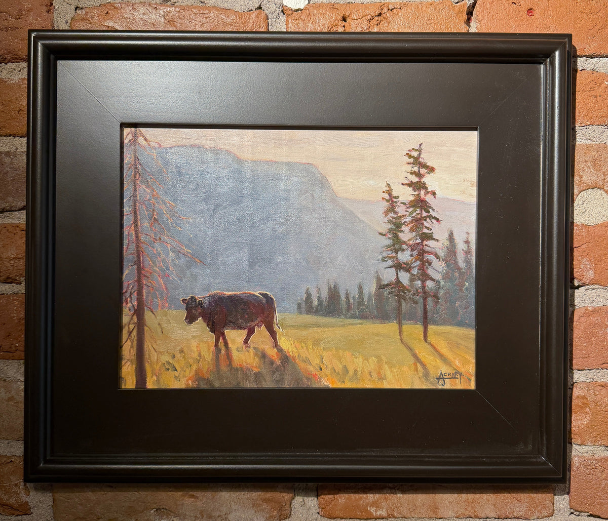 A framed landscape painting of a cow in a grassy field with mountains and trees, displayed on a red brick wall.