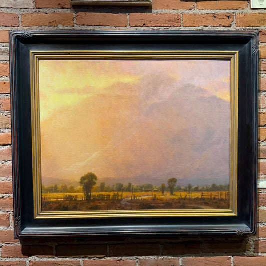 Painting of a landscape with trees and sky in a gold frame on a brick wall.