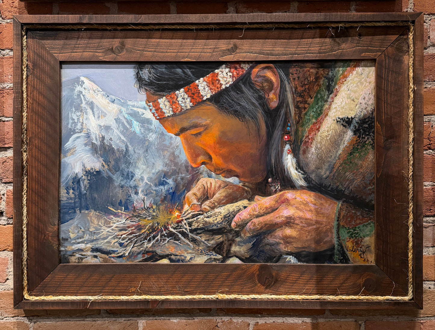Painting of a person preparing food with mountains in the background, framed and displayed on a brick wall.