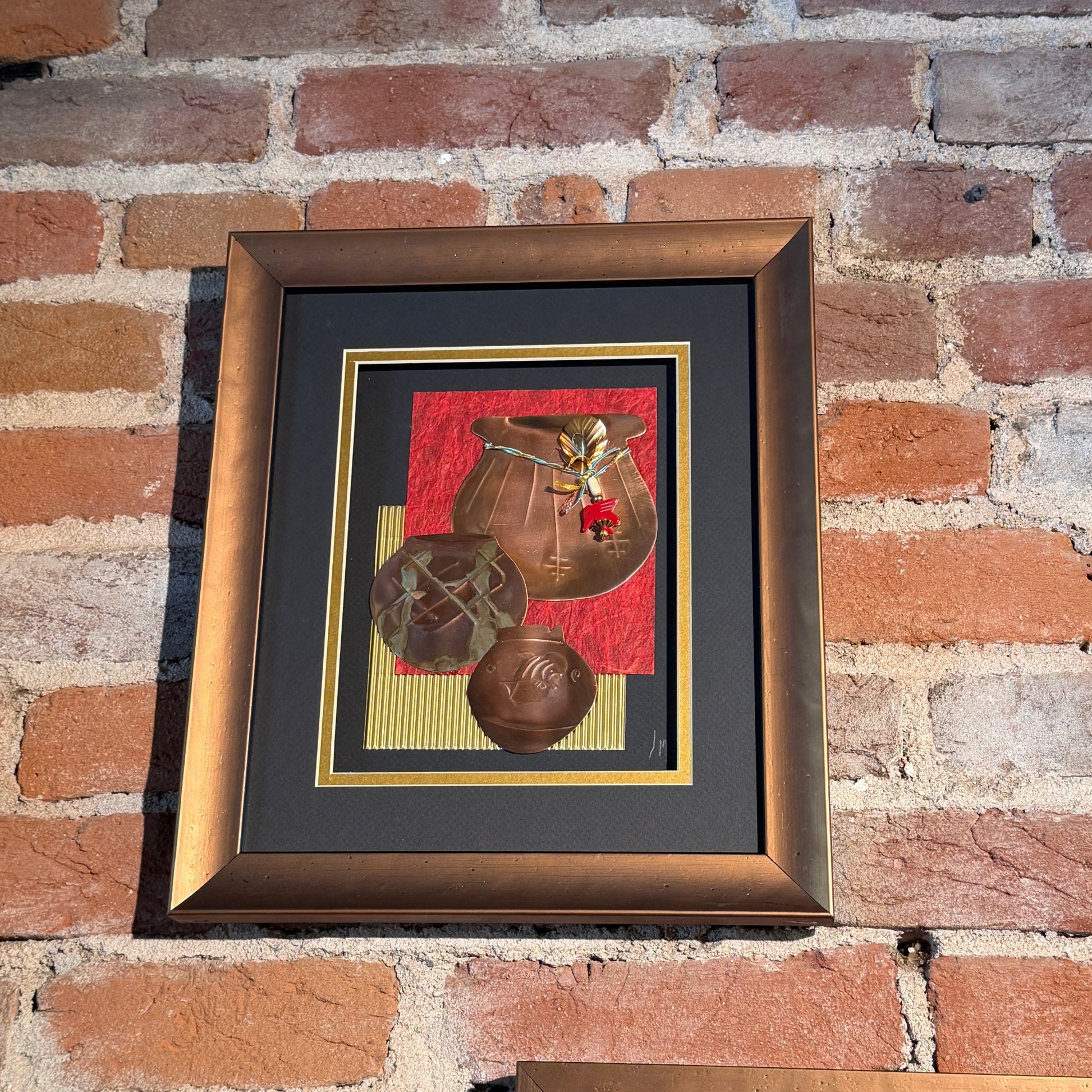 An art piece that contains thinly pressed copper shaped and detailed to represent three traditional Native American ceramic pots. They are placed in the foreground. The background is black gold, and red. Framed in a copper-colored frame