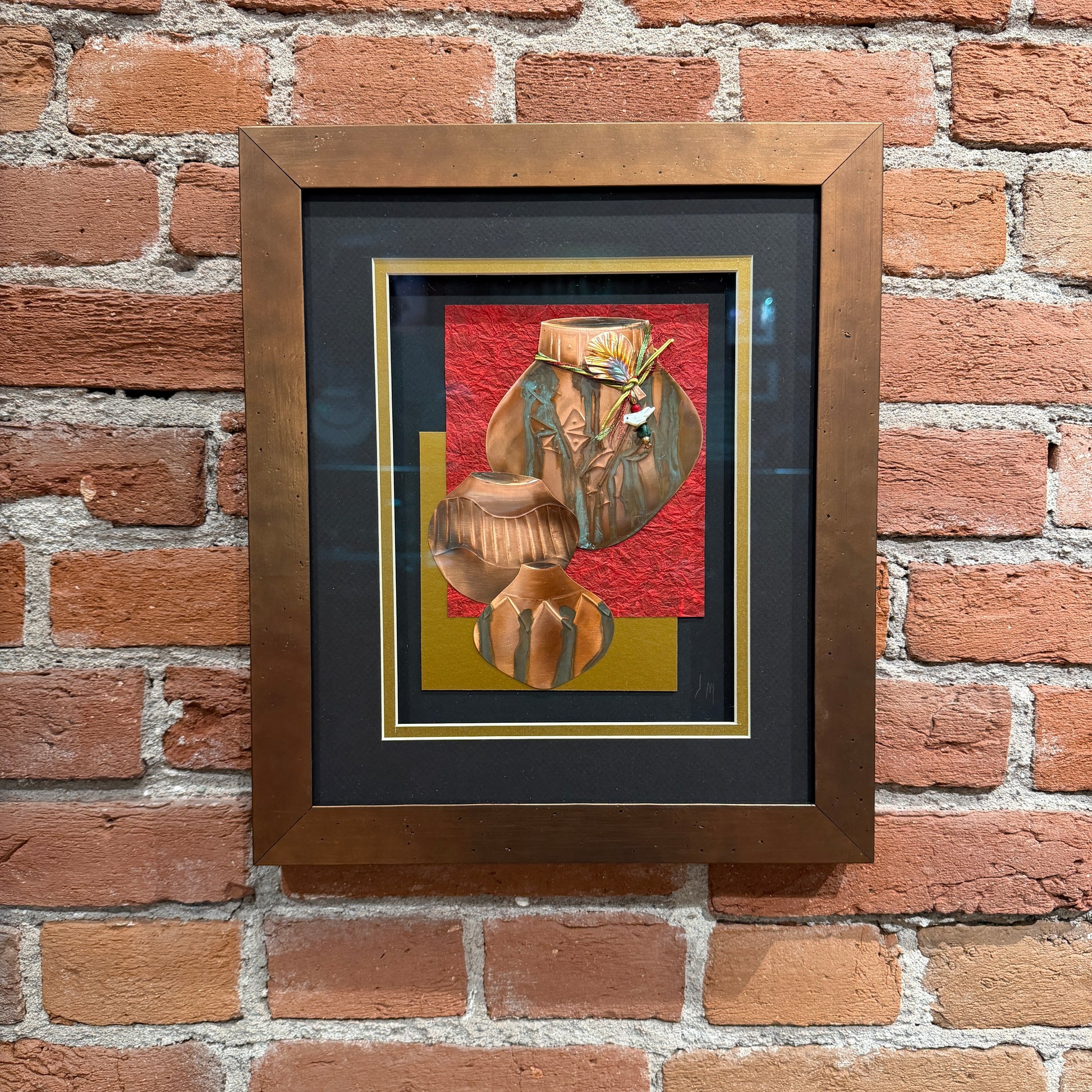 An art piece that contains thinly pressed copper shaped and detailed to represent three traditional Native American ceramic pots. They are placed in the foreground. The background is a black, gold and red. Framed in a copper-colored frame.