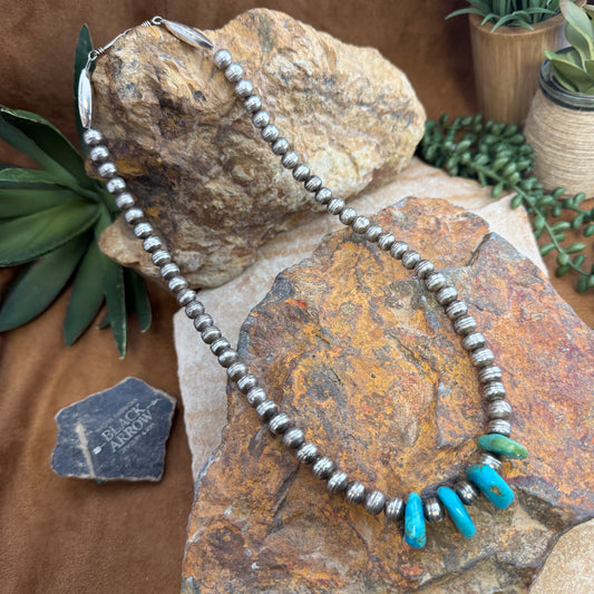 Silver beaded necklace with turquoise accents on a stone surface with plants