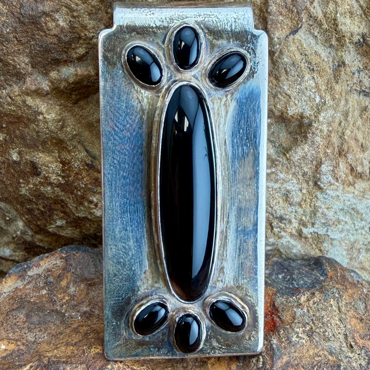 A rectangular silver-toned metal money clip featuring five glossy black stone inlays, photographed against a textured natural rock background.