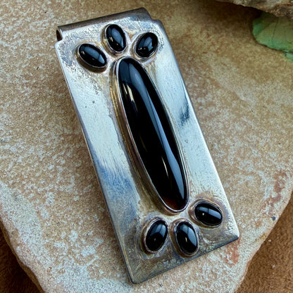 A handmade sterling silver money clip, featuring a central elongated oval inlaid with black onyx and four smaller oval accents around the border, resting on a textured stone surface.