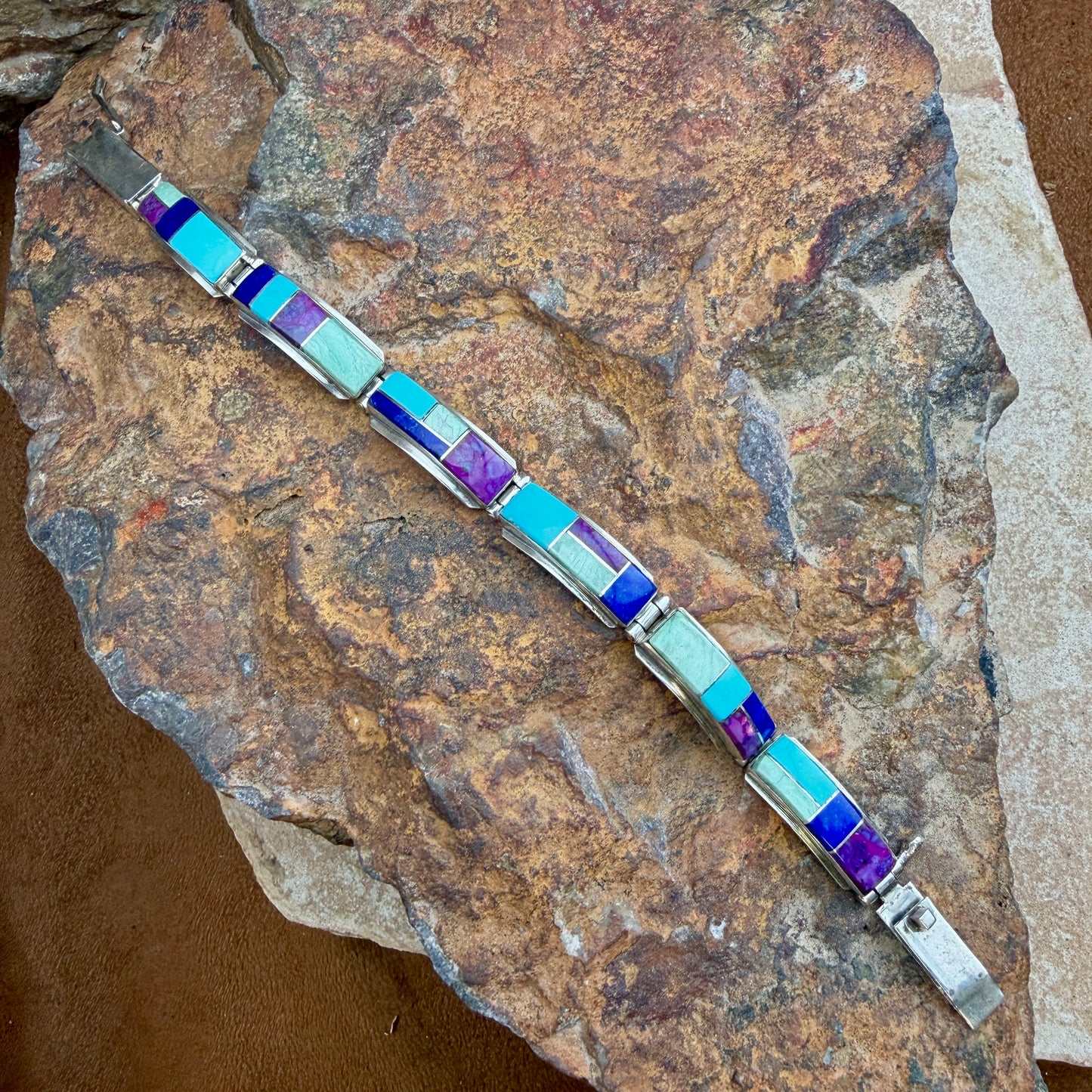 A silver-toned bracelet with rectangular inlay stones in light blue, purple, and pale mint, displayed diagonally on a textured brown rock.