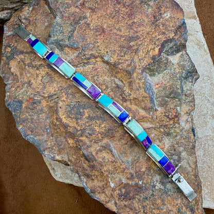 A silver-toned bracelet with rectangular inlay stones in light blue, purple, and pale mint, displayed diagonally on a textured brown rock.