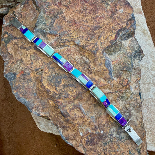 A silver-toned bracelet with rectangular inlay stones in light blue, purple, and pale mint, displayed diagonally on a textured brown rock.
