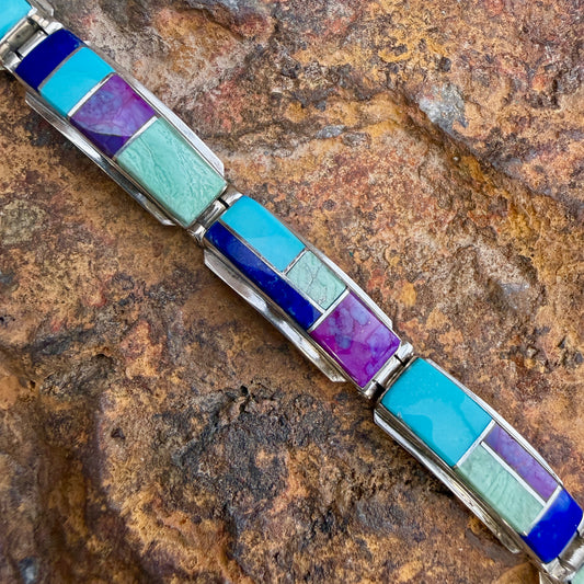 A silver-toned link bracelet featuring rectangular inlay stones in shades of turquoise, deep blue, purple, and pale mint green, displayed diagonally on a textured brown rock surface.