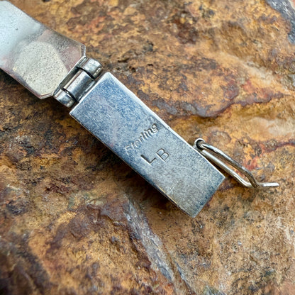 A close-up of a silver metal bracelet ending with a small attached ring, resting on a textured brown and tan rock surface. The metal is engraved with the word 'Sterling' and the initials 'LB'.