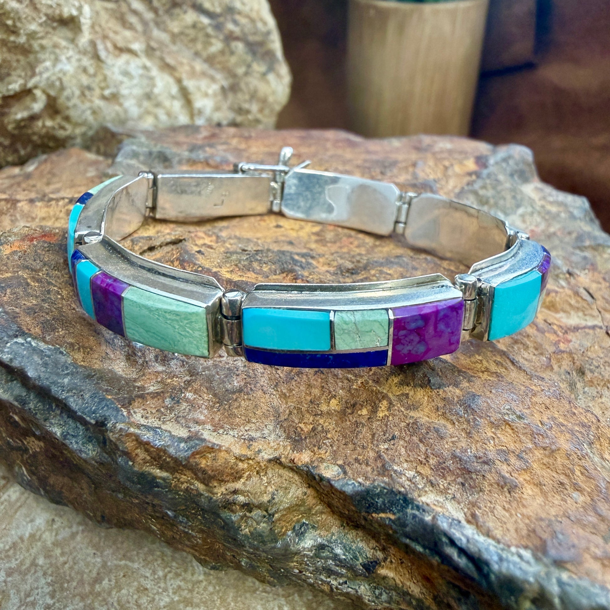 A silver-toned bracelet with rectangular turquoise, purple, and mint green inlay stones, displayed on a textured brown rock.