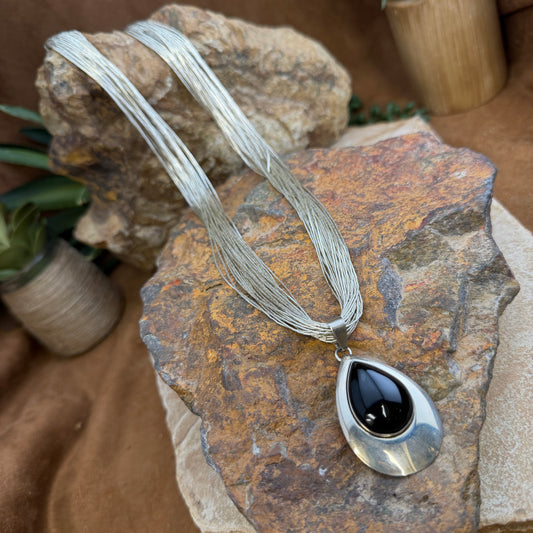 A silver-toned multi-strand necklace with a teardrop pendant featuring a glossy black center, displayed on textured brown and tan rocks.