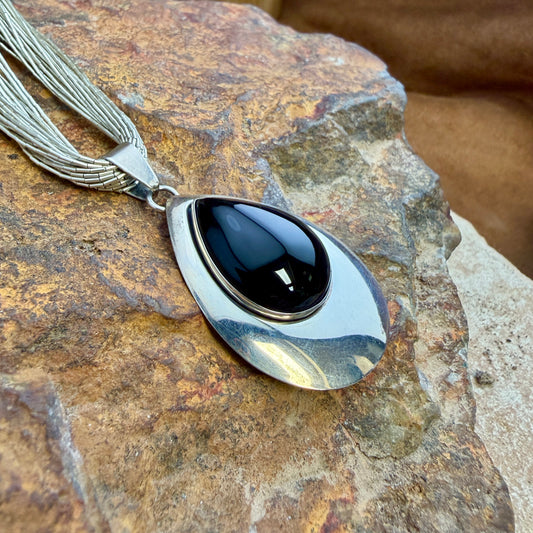 A silver-toned teardrop pendant necklace with a glossy black cabochon stone, resting on a textured brown rock.