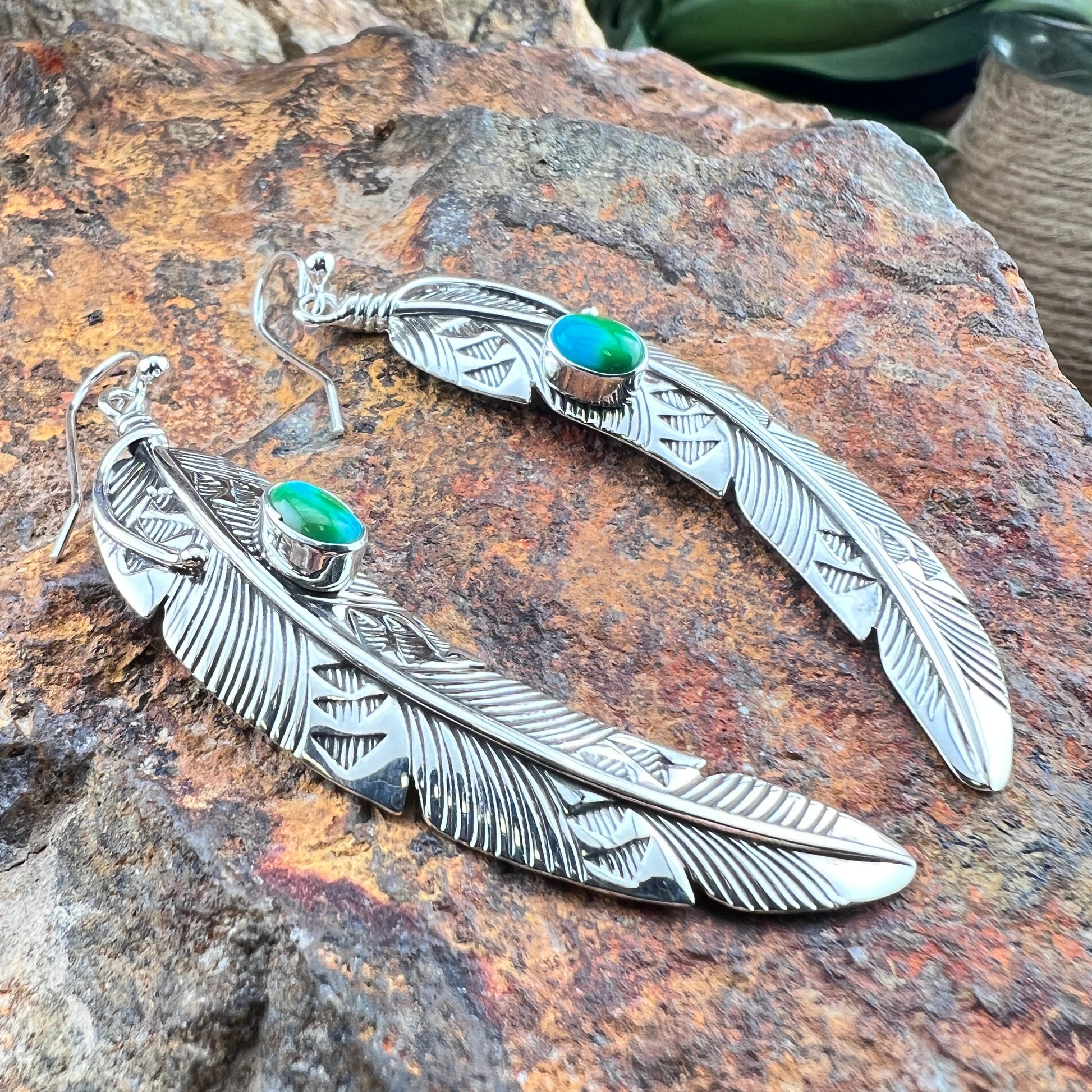 Silver feather-shaped earrings with green gemstones on a textured stone surface