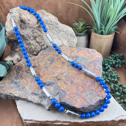 Blue beaded necklace with silver accents on a textured stone surface with plants in the background