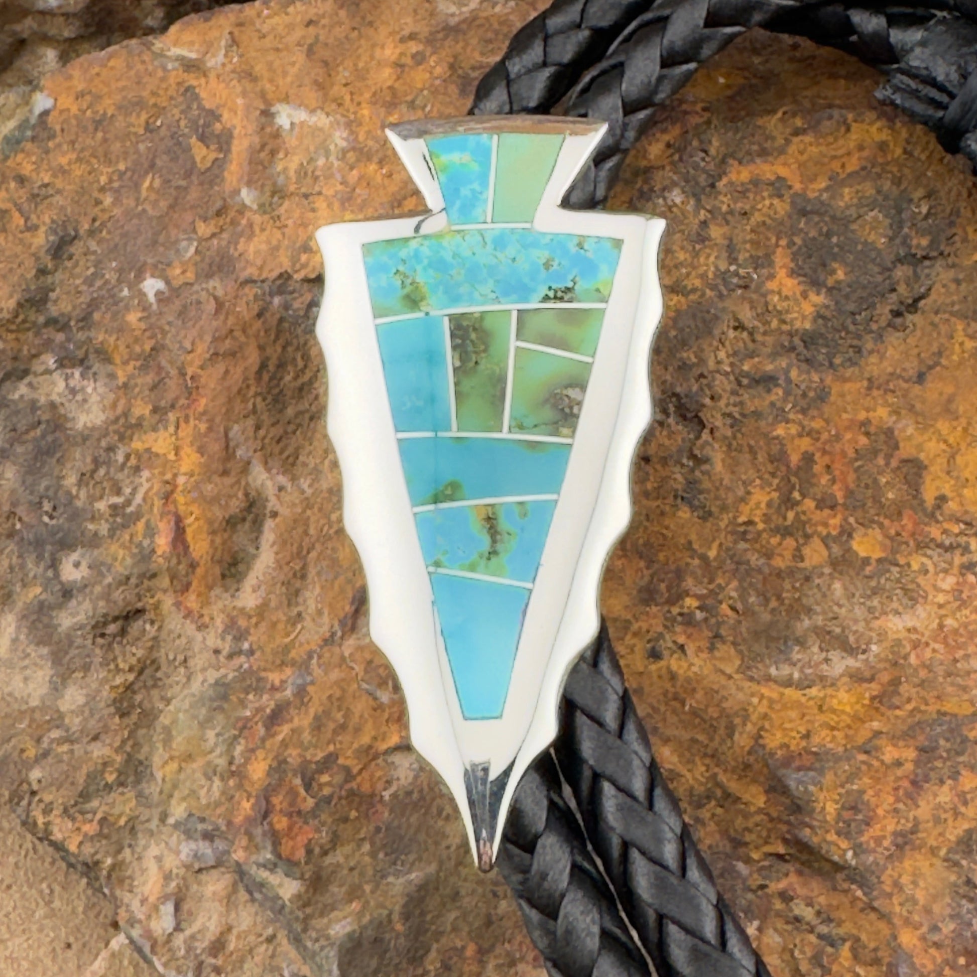 Arrowhead-shaped pendant with turquoise stones on a black braided cord against a stone background