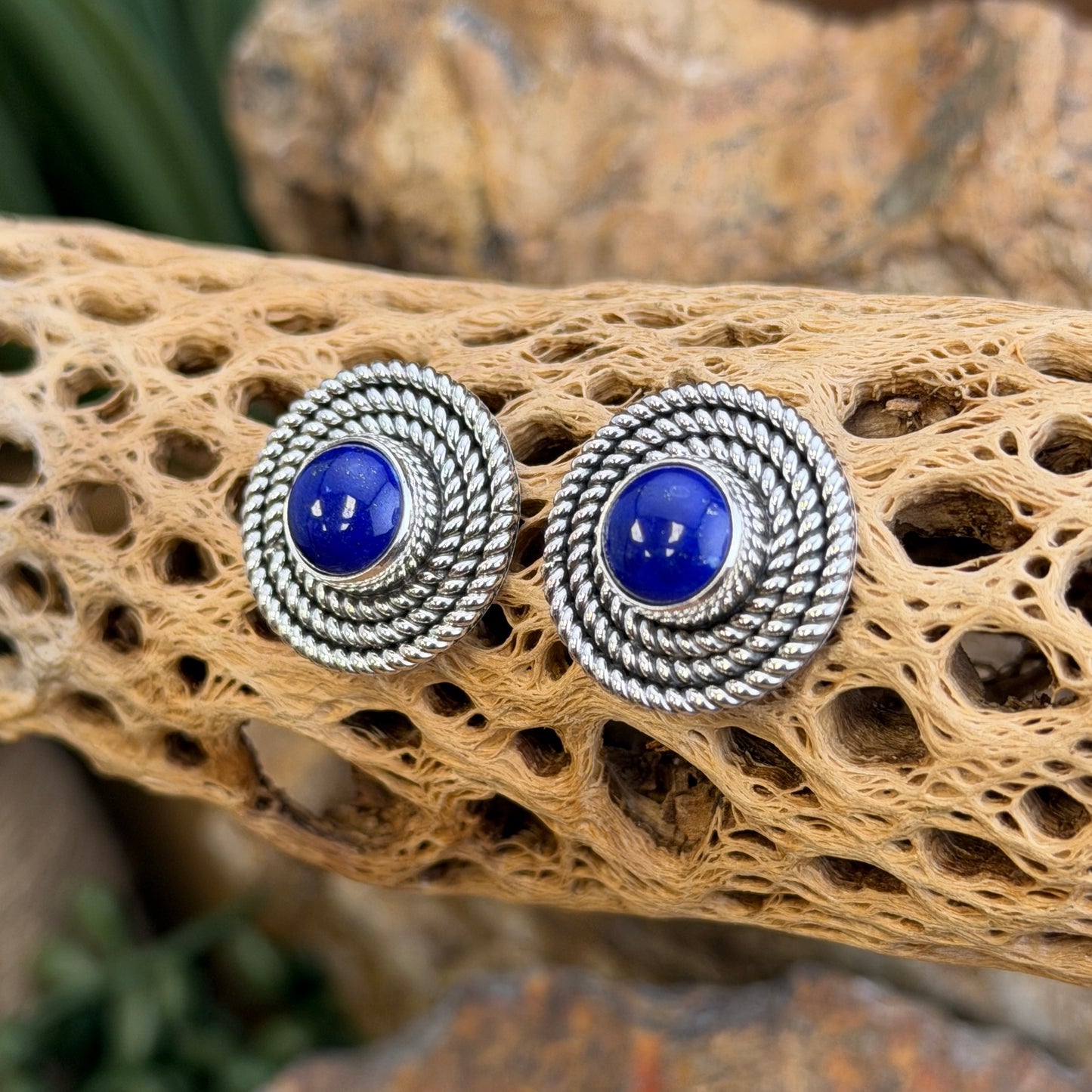 A pair of round stud earrings with deep blue cabochon stones and textured silver-toned rope-like borders, displayed on a natural tan driftwood piece with a blurred outdoor background.