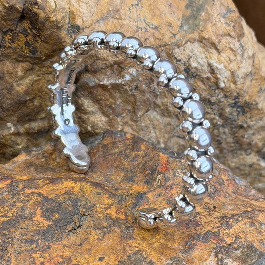 A polished silver cuff bracelet with a row of raised round beads and a textured open end, photographed resting on a textured brown and orange rock surface.