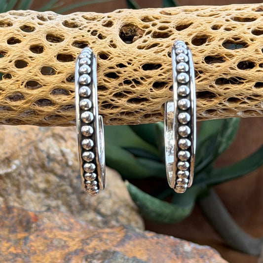 Two silver hoop earrings with bead detailing, displayed on a textured brown stone surface with a blurred natural background.