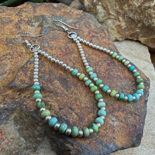 A pair of dangling beaded earrings with green and turquoise-blue round beads and silver-toned metal links, displayed on a textured brown rock outdoors.