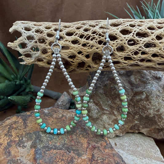 A pair of handmade teardrop hoop earrings with silver-tone metal and turquoise, mint green, and blue beads, displayed on a piece of driftwood over rocks with succulents in the background.