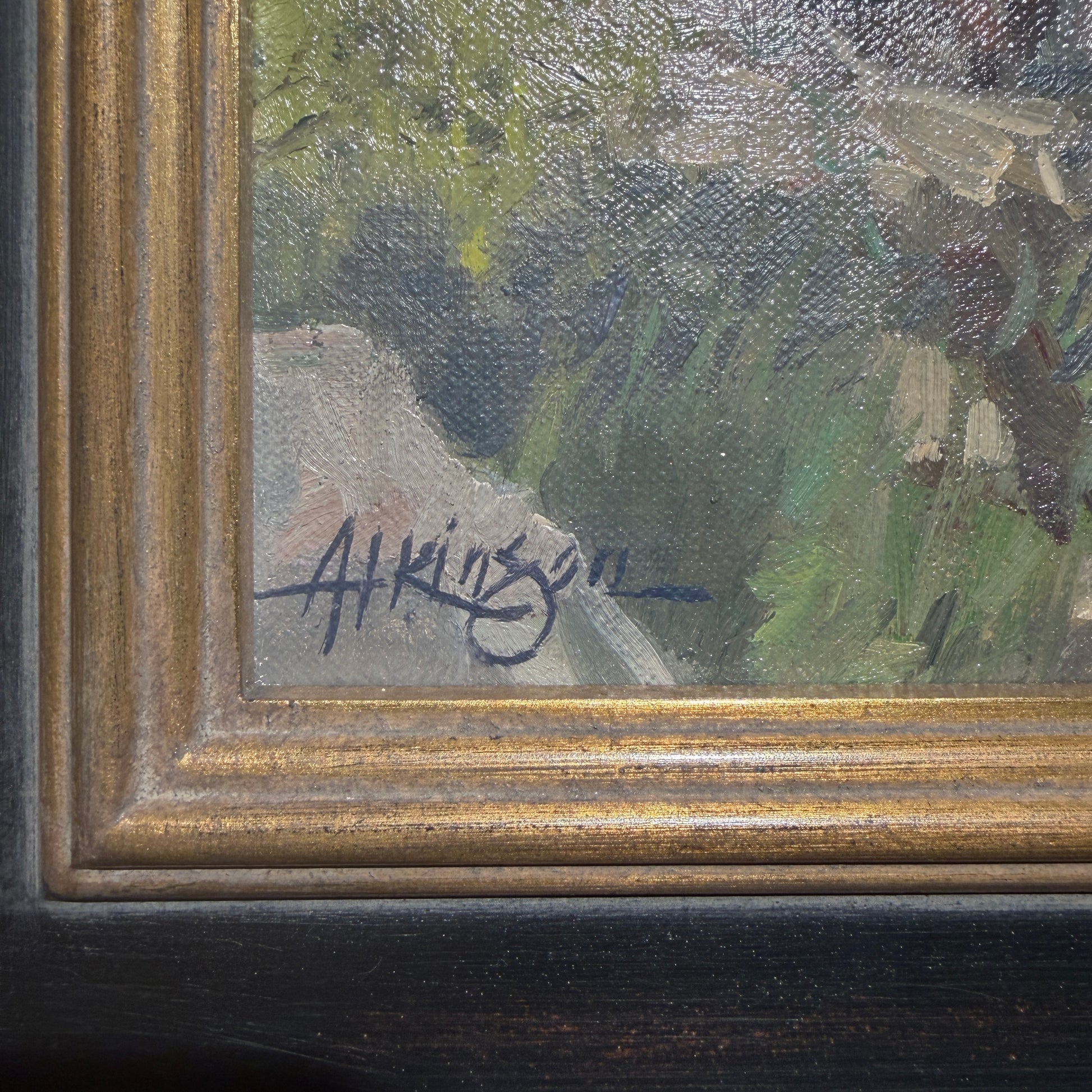 Close-up of a framed painting showing part of an outdoor landscape with green and gray brushstrokes, a gold-toned inner frame, and a dark wood outer frame. A handwritten signature is visible near the lower left corner.
