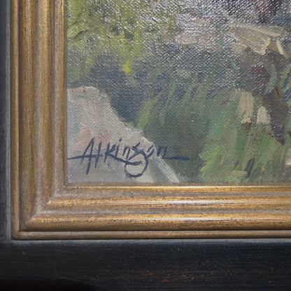 Close-up of a framed painting showing part of an outdoor landscape with green and gray brushstrokes, a gold-toned inner frame, and a dark wood outer frame. A handwritten signature is visible near the lower left corner.