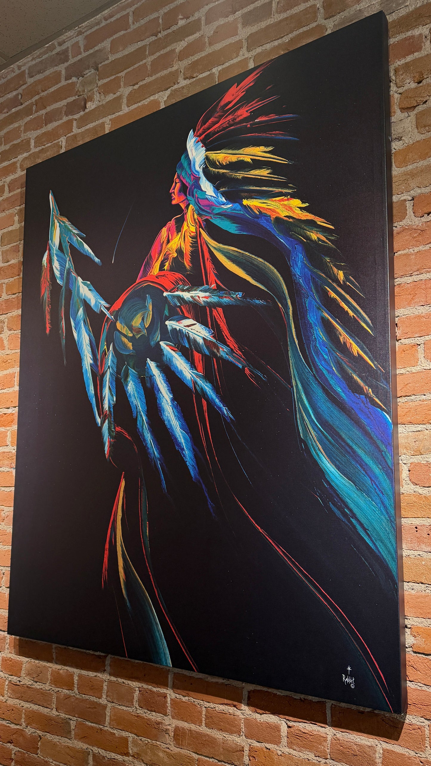 A large vertical canvas painting of a stylized warrior figure with a feathered headdress, rendered in vivid blue, red, yellow, and orange on a black background, mounted on a brick wall.