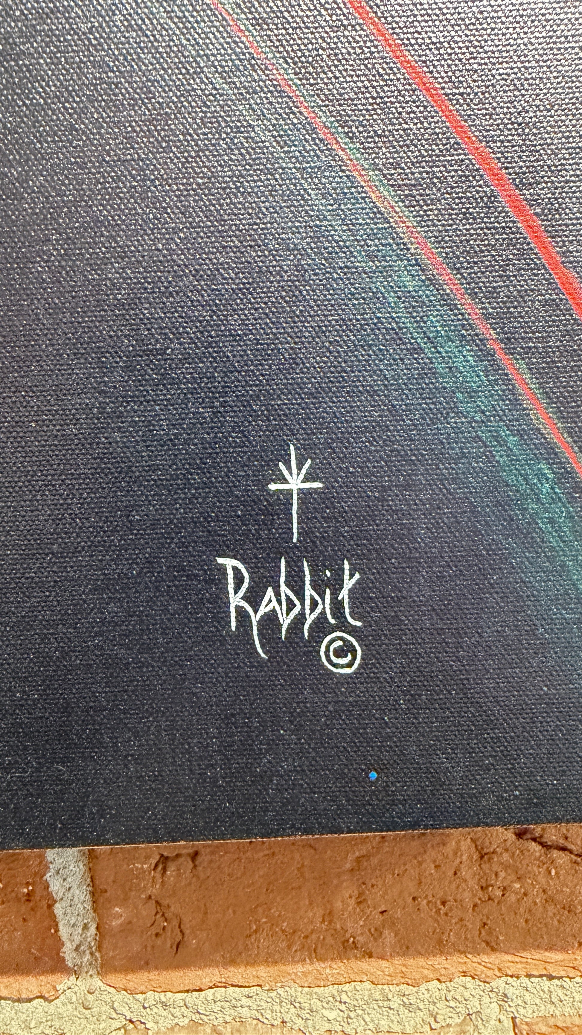 Close-up of a dark textured canvas or print with a white hand-drawn rabbit logo and circular mark near the center, plus thin red and teal curved lines in the upper right. A brick surface is visible along the bottom edge.