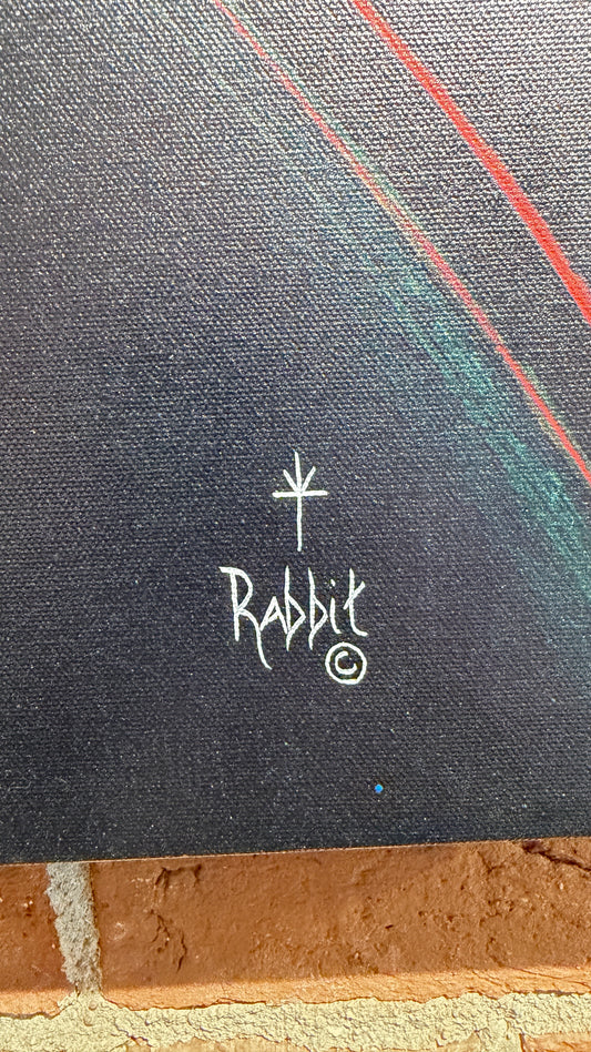 Close-up of a dark textured canvas or print with a white hand-drawn rabbit logo and circular mark near the center, plus thin red and teal curved lines in the upper right. A brick surface is visible along the bottom edge.