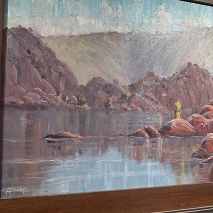 A framed landscape painting depicting a calm lake with reddish-brown rocky cliffs, reflections in the water, and a pale blue sky. The painting is shown at an angle with a dark wood frame visible at the bottom and left edge.