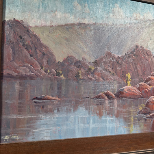 A framed landscape painting depicting a calm lake with reddish-brown rocky cliffs, reflections in the water, and a pale blue sky. The painting is shown at an angle with a dark wood frame visible at the bottom and left edge.