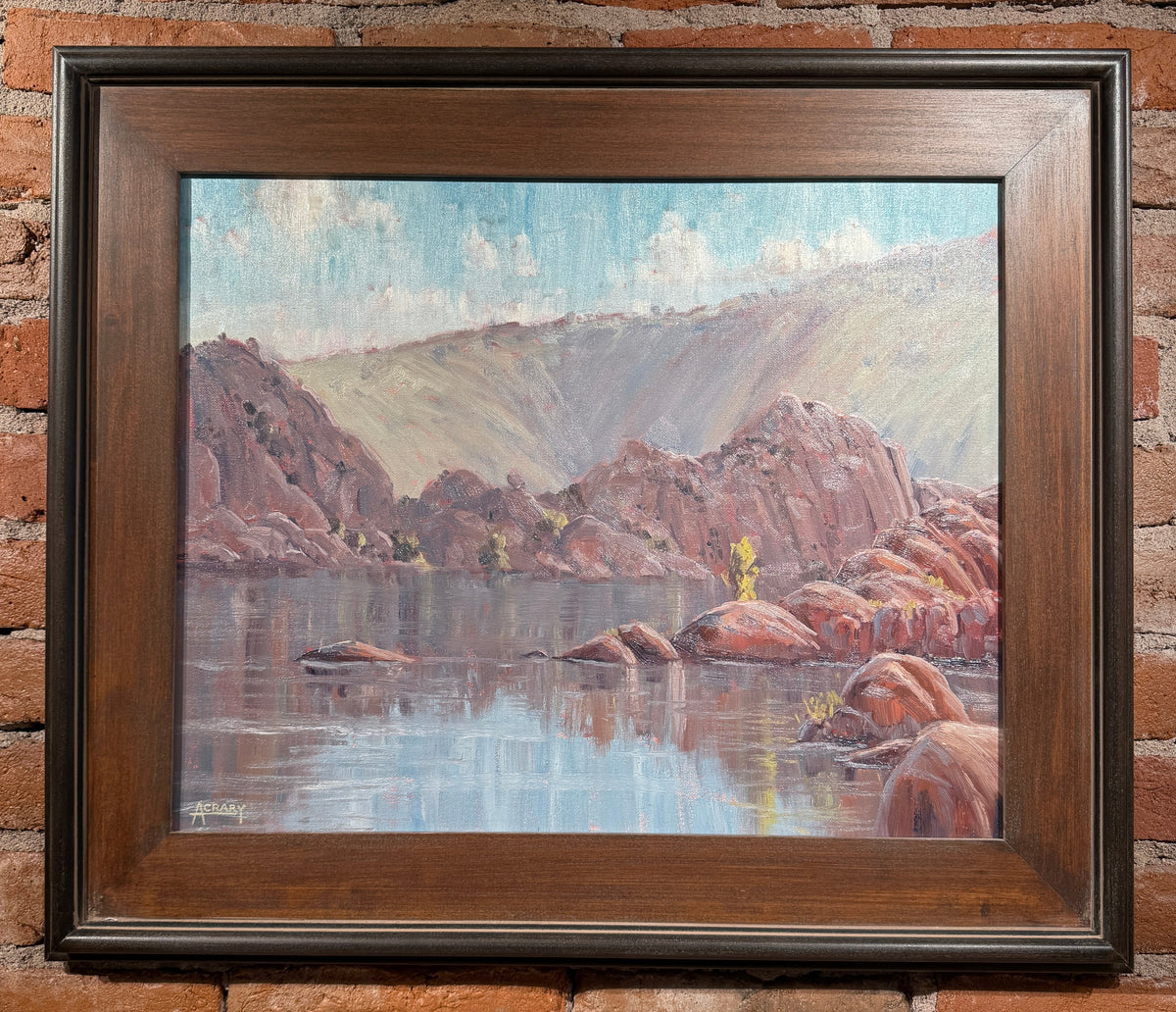A framed landscape oil painting depicting rocky red cliffs, a calm reflective body of water, and distant hills under a pale sky, mounted on a red brick wall.