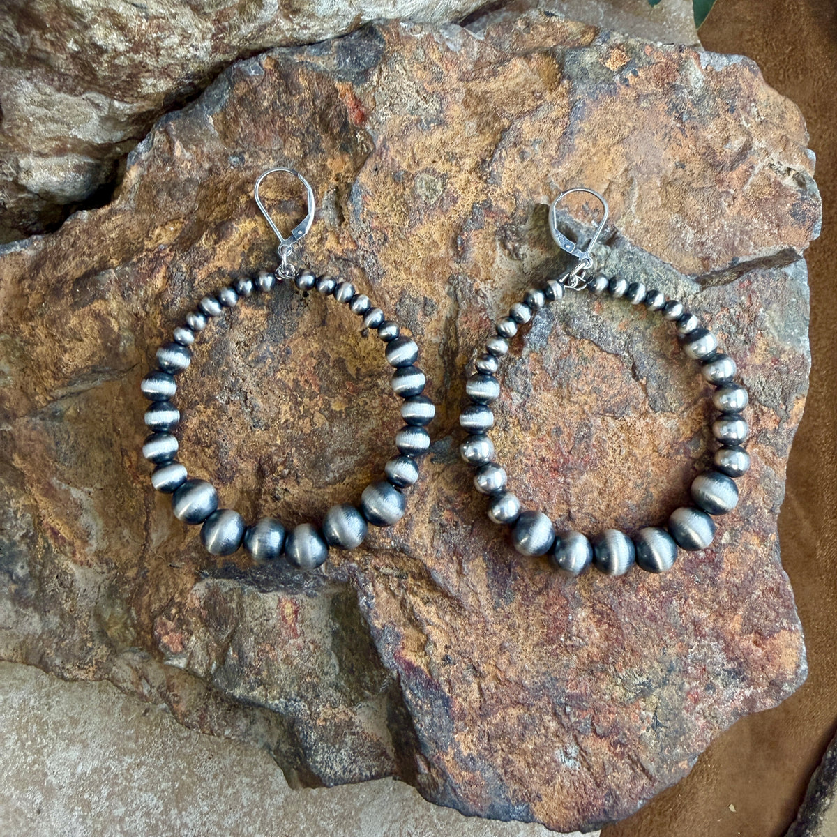 A pair of large hoop earrings made of Navajo pearls with a silver-toned fishhook back, displayed on a textured brown rock.