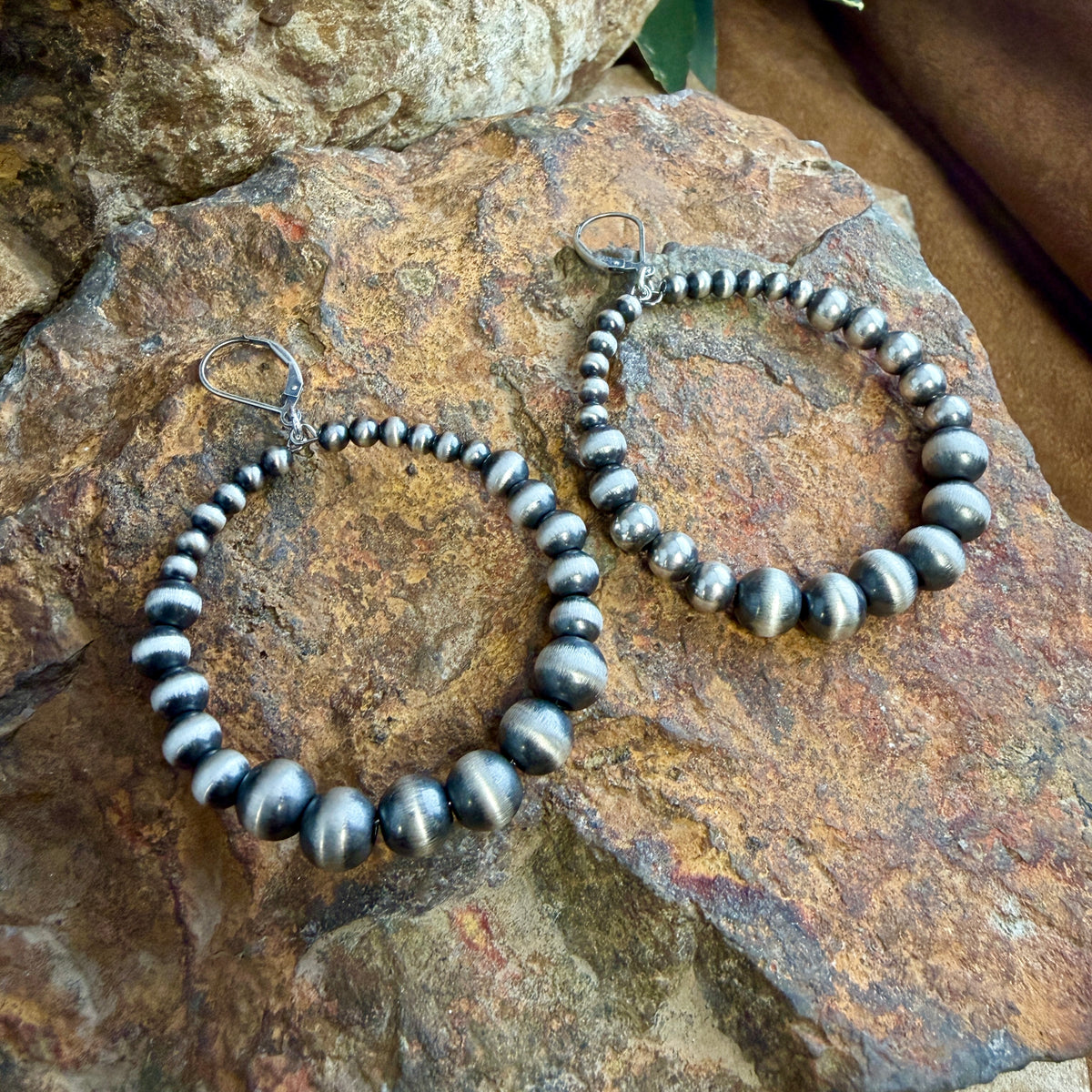 A pair of large sterling hoop earrings made of polished Navajo pearl beads, displayed on a natural rock.