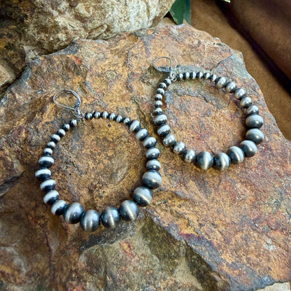 A pair of large sterling hoop earrings made of polished Navajo pearl beads, displayed on a natural rock.