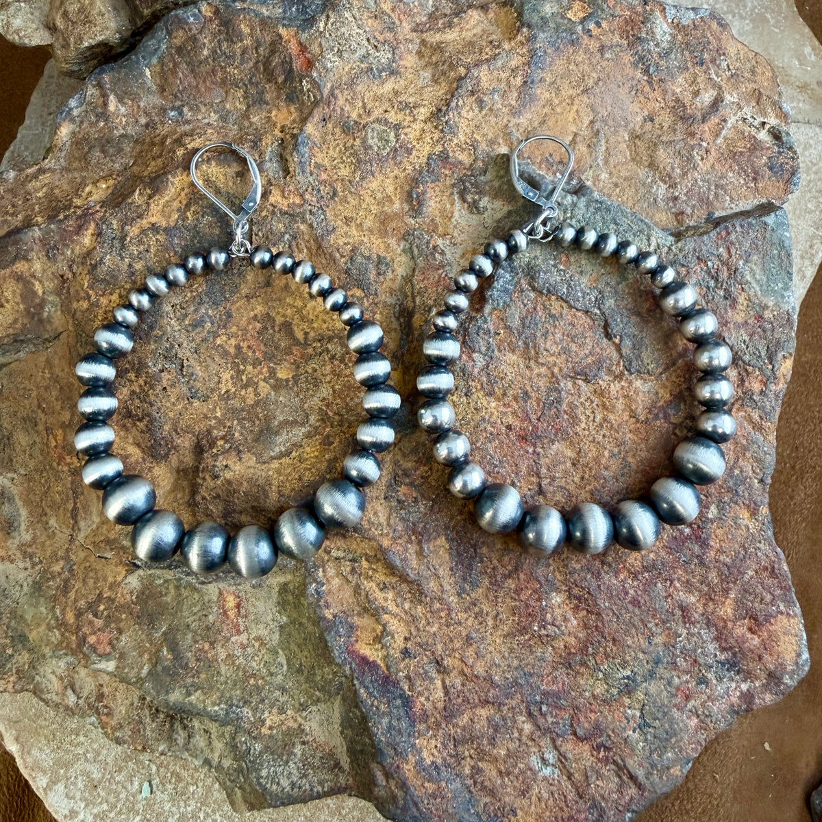 A pair of large sterling hoop earrings made of polished Navajo pearl beads, displayed on a natural rock.