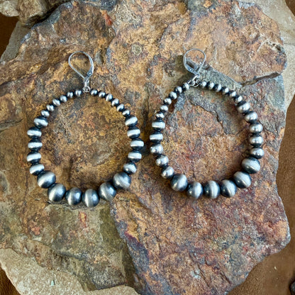 A pair of large sterling hoop earrings made of polished Navajo pearl beads, displayed on a natural rock.