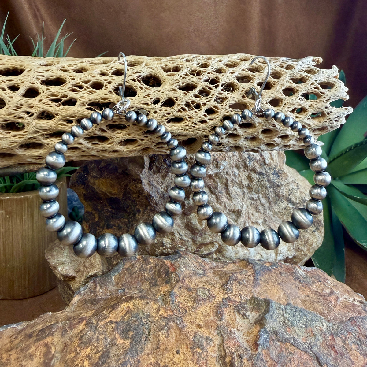 A pair of large silver-toned hoop earrings made of polished round beads, displayed on a natural rock and cactus bone with green plants in the background.