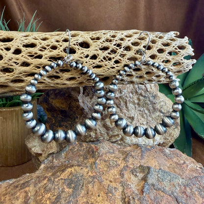 A pair of large silver-toned hoop earrings made of polished round beads, displayed on a natural rock and cactus bone with green plants in the background.