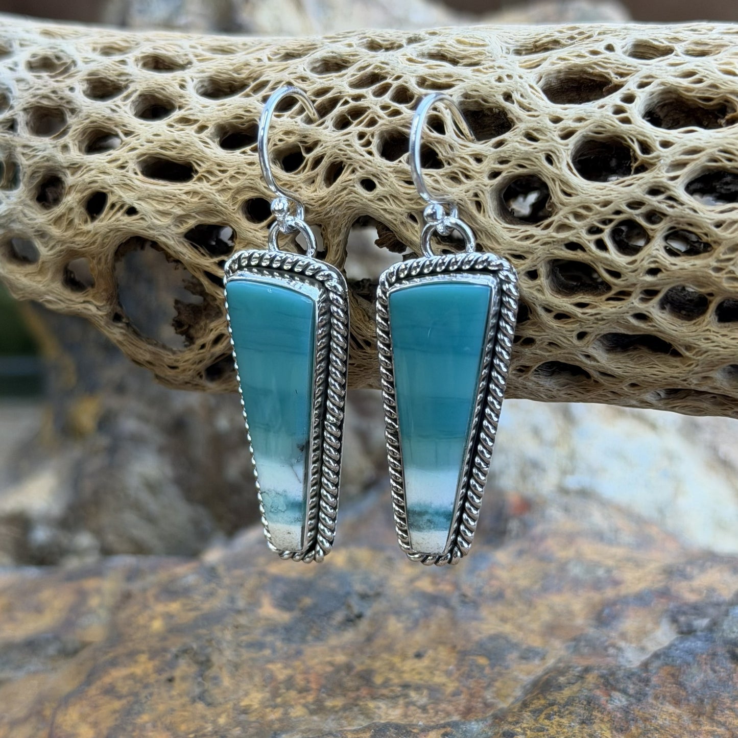 A pair of silver dangle earrings with teardrop-shaped turquoise-blue gradient stones, displayed on a natural cork log with a blurred rocky background.