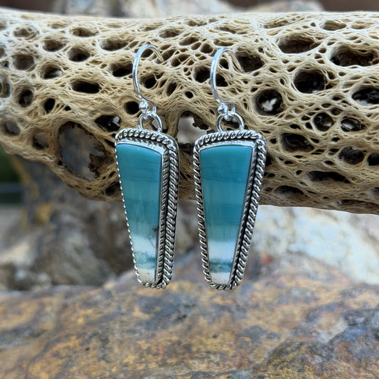 A pair of silver dangle earrings with teardrop-shaped turquoise-blue gradient stones, displayed on a natural cork log with a blurred rocky background.