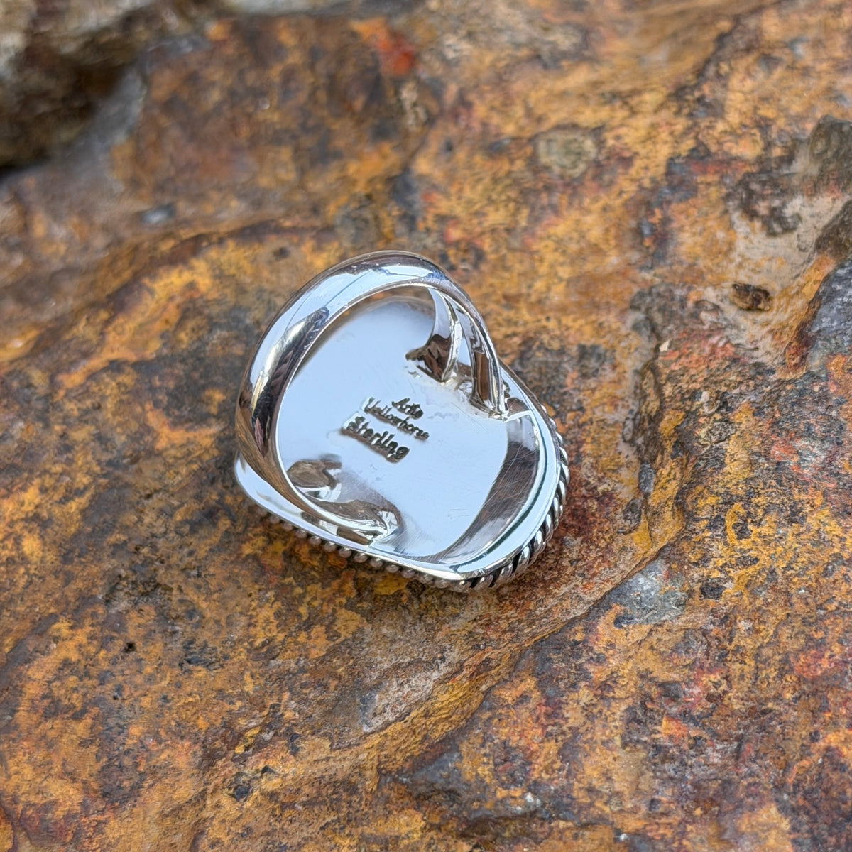 A Sterling Silver Ring with a large rectangular cabochon made of orange spiny oyster. it is surround by a double rope border. it rests on a rocky surface. this picture displays the back of the ring with "Artie Yellowhorse" and "Sterling"