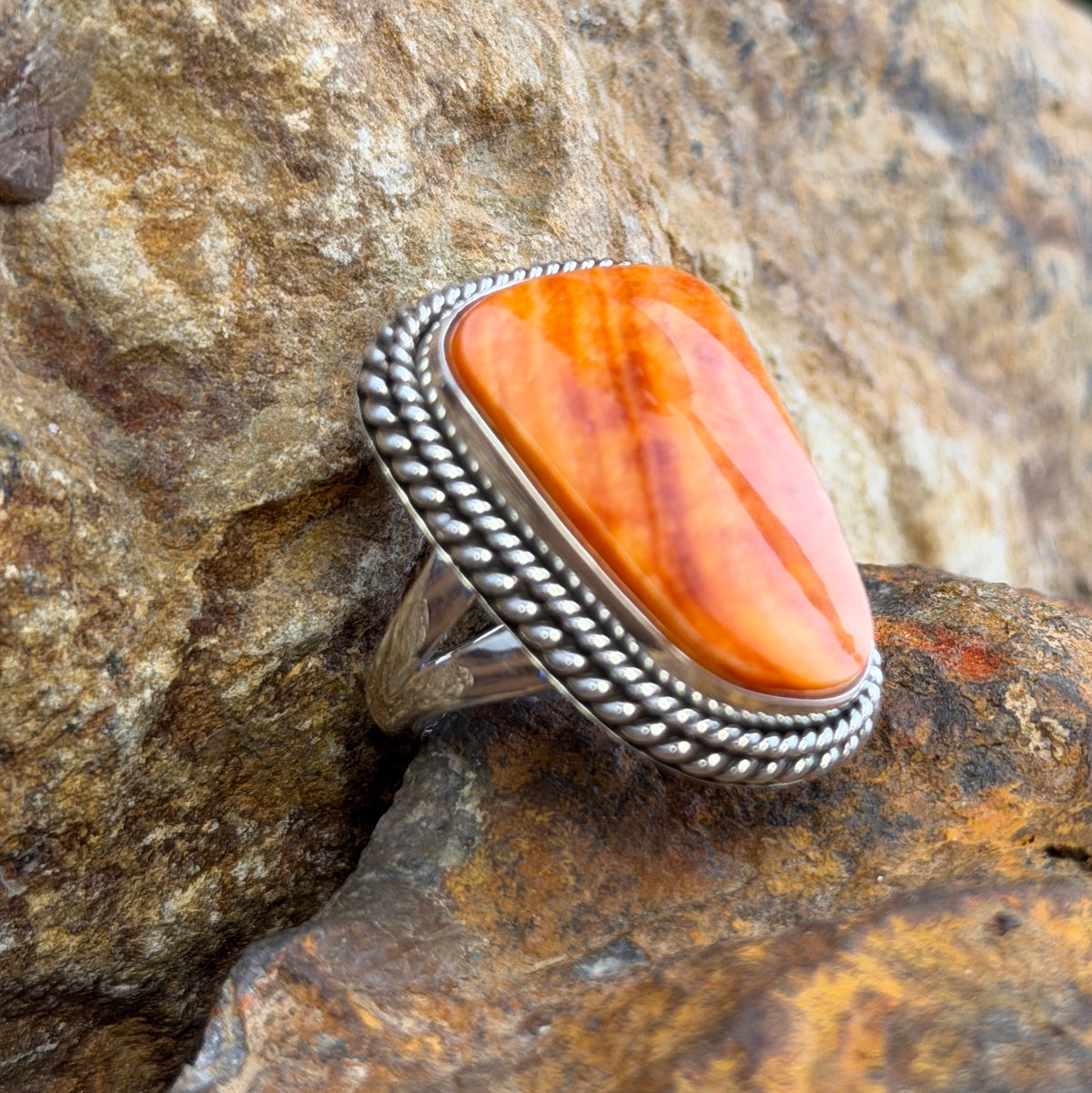 A Sterling Silver Ring with a large rectangular cabochon made of orange spiny oyster. it is surround by a double rope border. it rests on a rocky surface. 