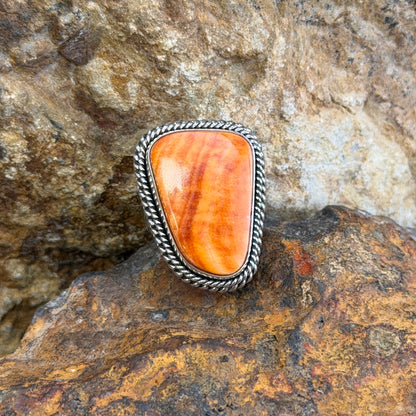 A Sterling Silver Ring with a large rectangular cabochon made of orange spiny oyster. it is surround by a double rope border. it rests on a rocky surface. 
