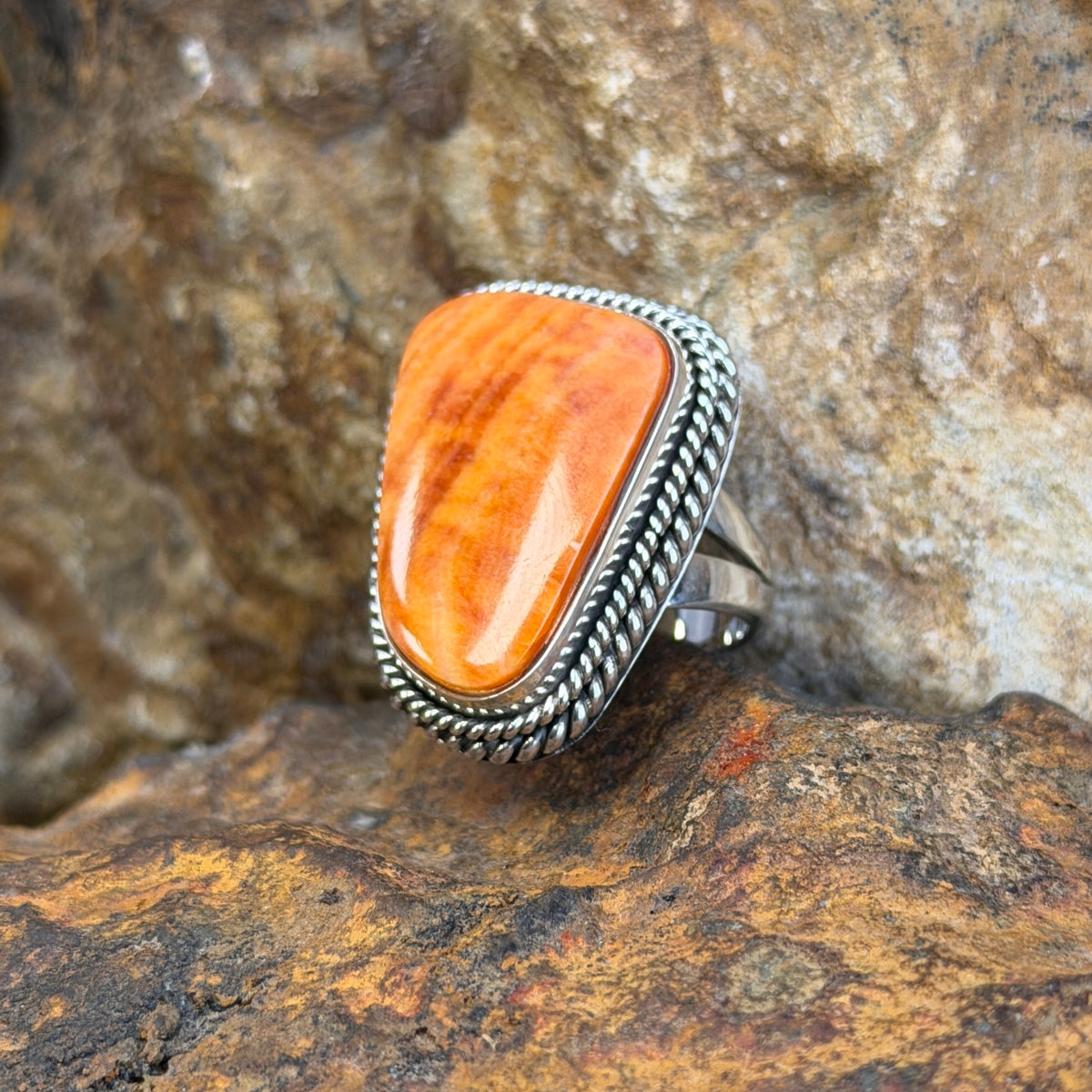 A Sterling Silver Ring with a large rectangular cabochon made of orange spiny oyster. it is surround by a double rope border. it rests on a rocky surface. 