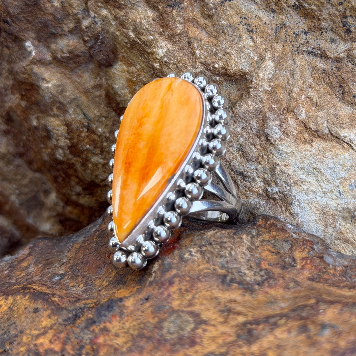 A sterling silver ring by Artie  Yellowhorse that holds an upside down teardrop cabachon made of orange spiny oyster. It is surrounded by silver beads. It rests on a rocky surface