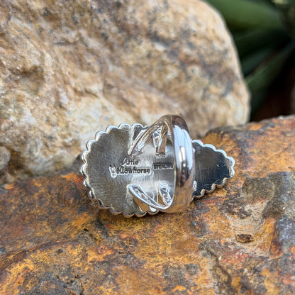 A sterling silver ring by Artie  Yellowhorse that holds an upside down teardrop cabachon made of orange spiny oyster. It is surrounded by silver beads. It rests on a rocky surface. This picture displays the inside of the ring to showcase the stamping of the artiest and "STERLING"